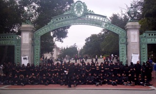 Hundreds of black students dressed in black, with faces covered, block a gate.